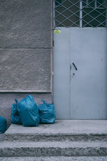 Four blue plastic rubbish bags are positioned on a concrete step in front of a grey metal door with a black handle, situated against a textured, weathered concrete wall. The scene appears to be outdoors, possibly on a building's entrance or service area, with the bags neatly gathered and leaning slightly to the left side of the door. The door features a small, rectangular yellow notice above the handle, and the upper section of the door includes a grid-patterned metal grate or window. The environment suggests a private area where rubbish is collected outside for either disposal or alternative waste collection, which may be managed by a service such as Rubbish Removal Belgravia, specializing in local waste management and rubbish removal services. The lighting is natural, highlighting the details and textures of the wall, door, and rubbish bags, with no visible activity or further context outside the immediate area of the rubbish bags and door.