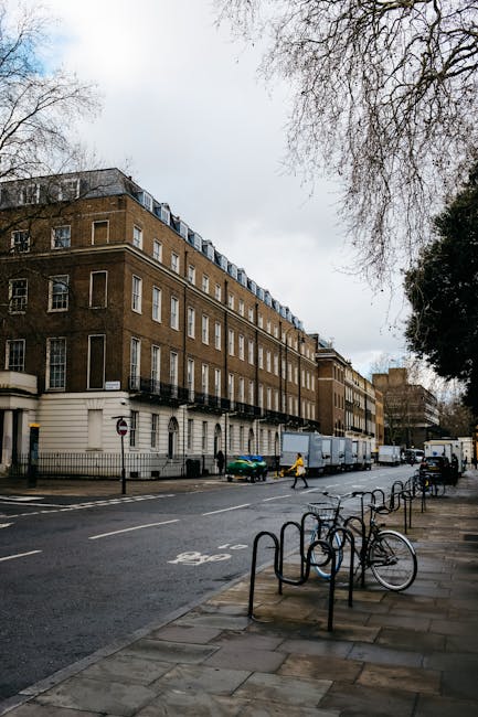 The image depicts a city street scene with a large, multi-storey brick building on the left side, featuring numerous evenly spaced rectangular windows and a cream-colored base. In the foreground, a paved sidewalk runs parallel to the road, which has a line of metal bike racks fixed to the pavement, some of which are empty while others have bicycles securely chained to them. The street itself is relatively empty, with only a few pedestrians walking along the sidewalk—including one person in a yellow jacket—and a small green car parked near the building. Further down the street, a white delivery truck is parked, partially blocking the view of more buildings in the background. Bare trees with leafless branches extend into the frame from the upper right corner, suggesting it is late autumn or winter. The scene is well lit with natural daylight, and the overall atmosphere is quiet and orderly, typical of an urban environment where private waste collection and rubbish removal services, such as those provided by Rubbish Removal Belgravia, could be involved in ongoing waste management or clearance projects in the area.