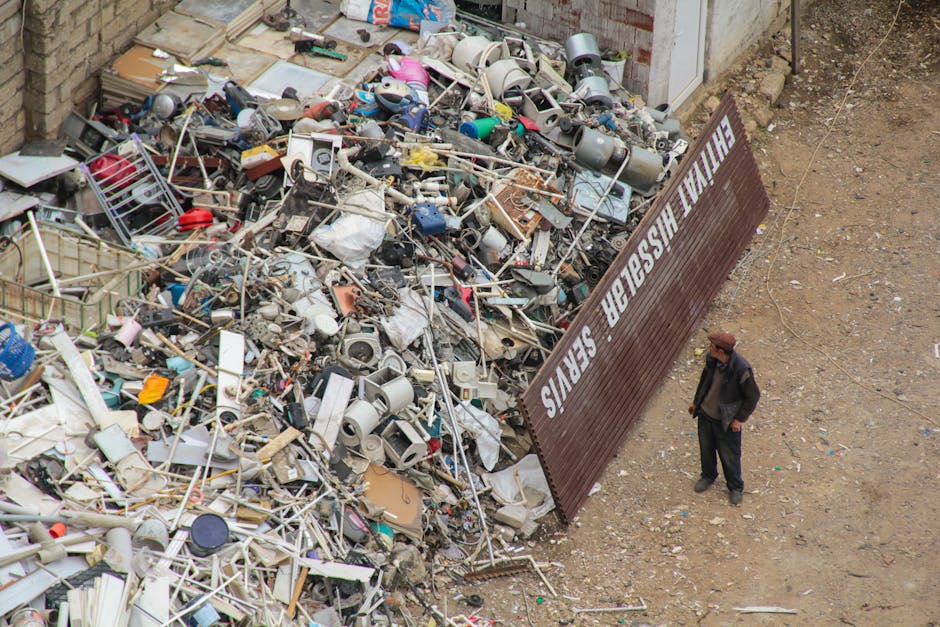 A large pile of mixed domestic waste, including broken electronic devices, plastic pipes, metal frames, and discarded household items, is seen outdoors on a gravel surface next to a brick wall. The debris appears to be a combination of broken furniture, appliances, and miscellaneous trash materials, with some items partially covered by paper or plastic wrapping. Leaning against the pile is a weathered, dark brown metal fence panel with partially obscured white lettering, reading 'THE MAIL H' and other letters not fully visible. A lone man, dressed in a dark jacket and trousers with a brown hat, stands nearby observing the rubbish. The environment suggests an outdoor waste disposal or collection site, possibly related to private rubbish removal services, with no indication of recycling or proper sorting in place. The scene is lit naturally, emphasizing the cluttered and unmanaged nature of the waste, indicative of an area requiring professional waste management, such as services provided by Rubbish Removal Belgravia.