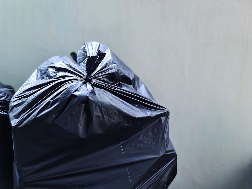 Close-up of several black garbage bags made from shiny, rippled plastic, stacked against a plain, light-colored wall. The bags appear filled and tied at the top with tight knots, with visible creases and folds in the plastic material. They are positioned in the left foreground, with one bag slightly leaning against another, suggesting recent collection or awaiting removal. The background shows a smooth, neutral-toned surface with minimal features, indicating an indoor or sheltered outdoor environment such as a storage area or alleyway for waste collection. The lighting is soft and diffuse, emphasizing the reflective quality of the plastic packaging. This scene exemplifies typical waste receptacles prepared for private rubbish removal services, aligning with the operational context of a company like Rubbish Removal Belgravia providing alternative waste management solutions for residential or commercial clients in the area.