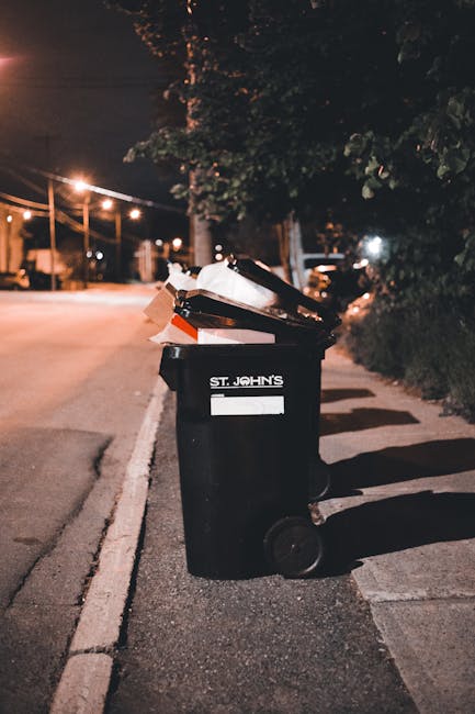 A black wheeled rubbish bin labeled 'St. John's' positioned on a residential street at night, with the lid partially open revealing waste materials inside. The bin is placed on the edge of a paved sidewalk, adjacent to a curb and a row of dense greenery or bushes to the right. In the background, there are illuminated streetlights casting a warm glow over the empty street and faint outlines of trees and parked vehicles. The scene captures an urban environment during late evening, highlighting the importance of proper waste collection as part of neighbourhood maintenance, with the bin ready for collection or disposal. The image reflects the routine of waste management services like those provided by Rubbish Removal Belgravia, emphasizing the practical aspects of rubbish removal and private waste handling within a UK residential context.
