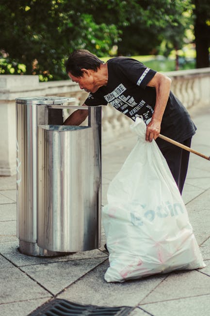 An elderly woman with short dark hair, dressed in a black T-shirt with white text on the front, is seen discarding rubbish into a stainless steel public litter bin located on a paved sidewalk. She is leaning forward slightly, holding a large white trash bag in her left hand, which appears to be filled with waste and partially draped over her arm. The woman's right hand is extended into the top opening of the bin, actively discarding waste. The bin has a modern cylindrical design with a smooth, reflective surface and a rectangular opening positioned at the top, on a sidewalk near a low stone wall. In the background, there are lush green trees and foliage, indicating a park or street with natural surroundings. The overall scene suggests a community effort in waste collection and litter management, underlining the importance of regular rubbish removal and street cleanliness, often managed by services like Rubbish Removal Belgravia for alternative waste handling outside of council collections.