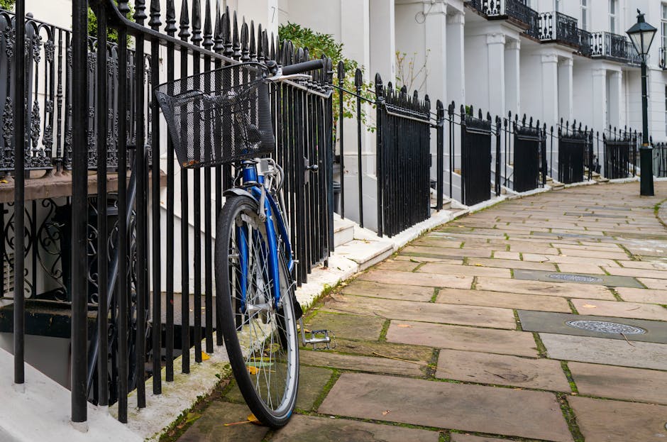 In the foreground, a blue bicycle with a black front basket is locked to a black metal railing along a pavement in a residential area. The railing features vertical bars with decorative finials at the top, running parallel to the white-painted building facades behind it. The building has white columns and black iron balconies, indicating classic architectural details typical of a London terrace. The pavement is paved with large, irregularly shaped stone slabs, showing slight moss or grass growth between the joints, and curves gently along the street. The scene is evenly lit with natural daylight, highlighting the textures and colors of the materials—metal, stone, and painted surfaces. To the right, a series of small black bollards separates the pedestrian walkway from the street. The environment appears clean and well-maintained, reflecting the typical setting where private rubbish collection and on-site clearance services by Rubbish Removal Belgravia could be utilized for waste management or property clearing in the area, although no rubbish or disposal objects are visible in this scene.